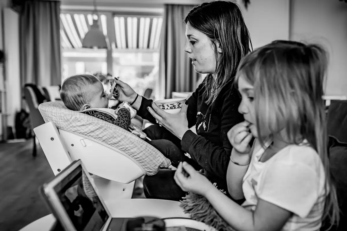 Moeder voed baby terwijl oudste dochter achter iPad eet.