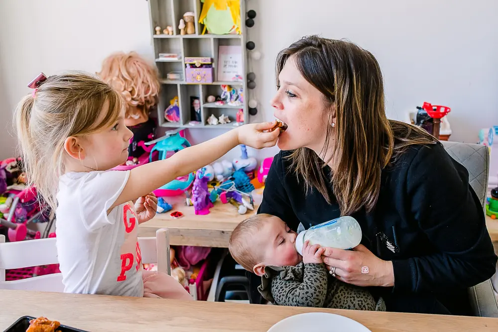 Oudste dochter geeft moeder die een baby een fles geeft een hapje.