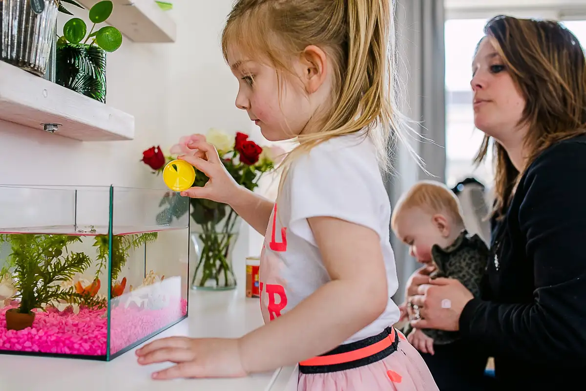 Meisje geeft visjes in aquarium eten terwijl moeder toekijkt.