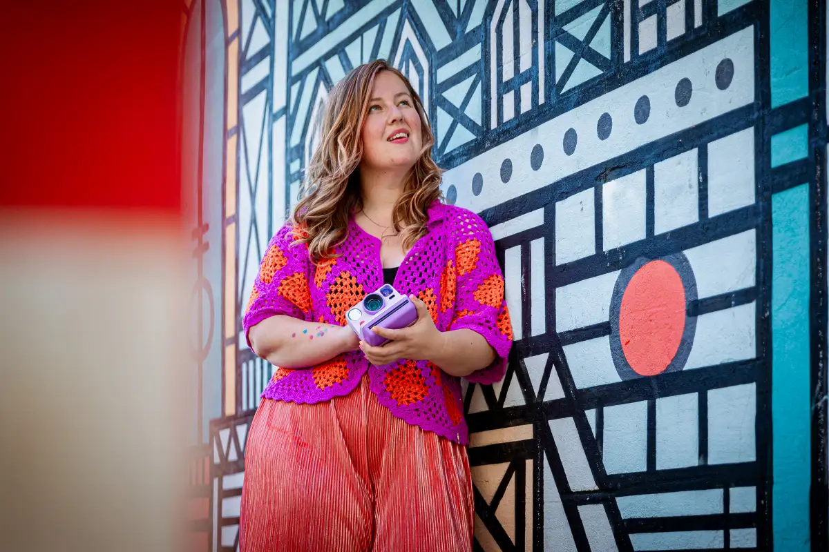 Vrouw poseert met lila polaroid camera bij een beschilderde muur in Breda.