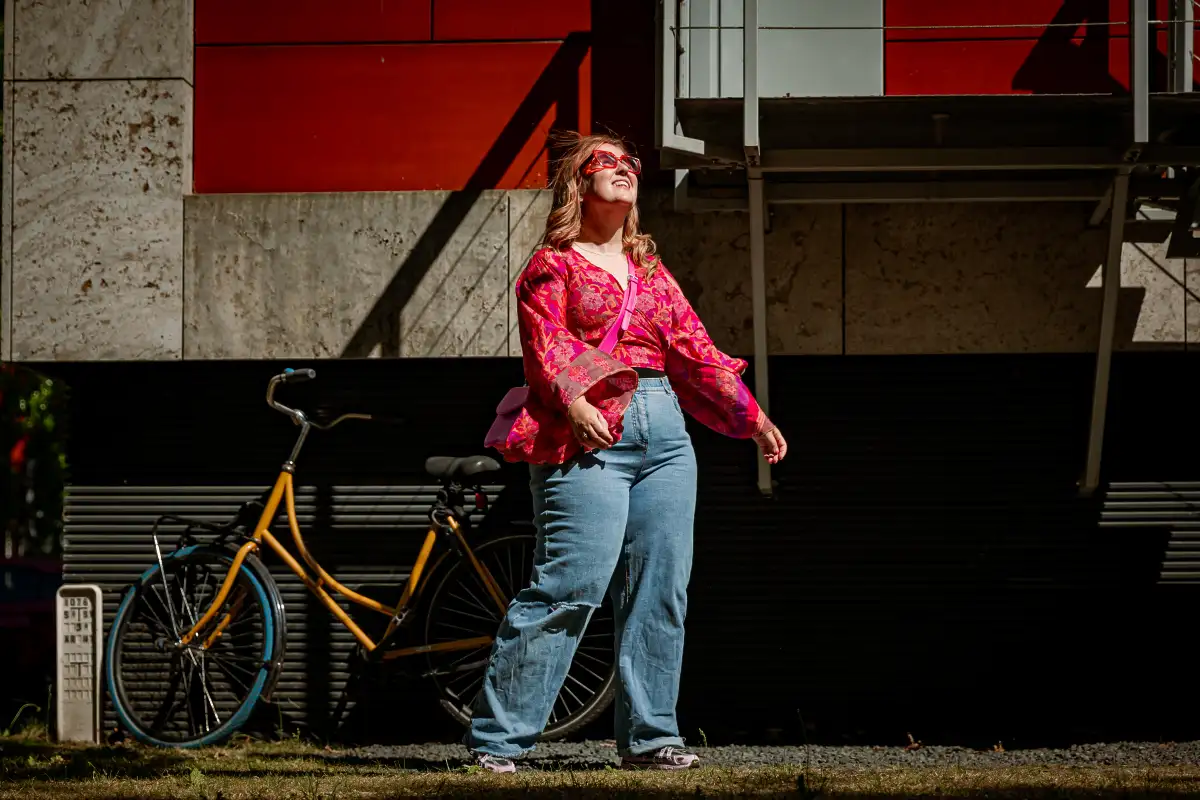 Vrouw bij woning met gele fiets kijkt met zonnebril richting de zon.