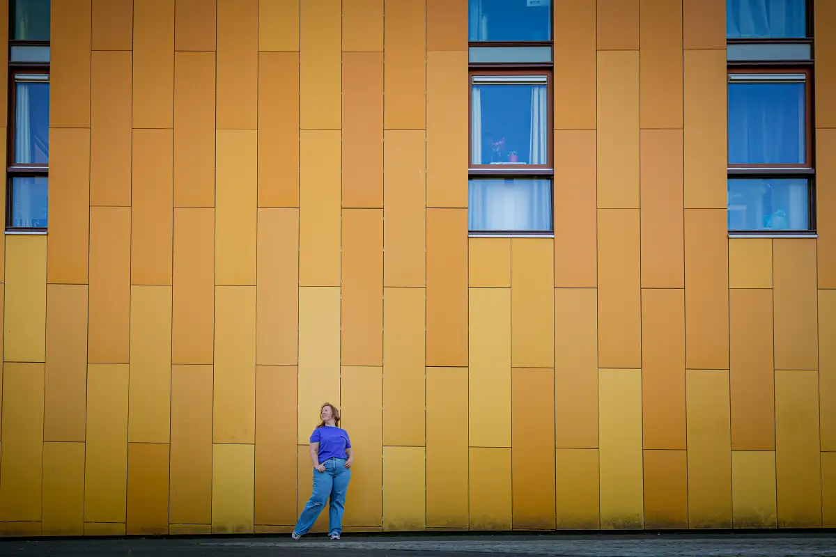 Vrouw in paars shirt en spijkerbroek poseert bij gele muur bij Chassé Promenade in Breda.