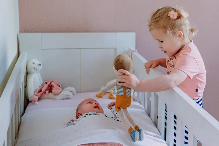Grote zus laat een pop zien aan baby in bedje.