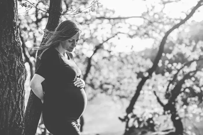 Zwangere vrouw leunt tegen boom en kijkt naar haar buik.
