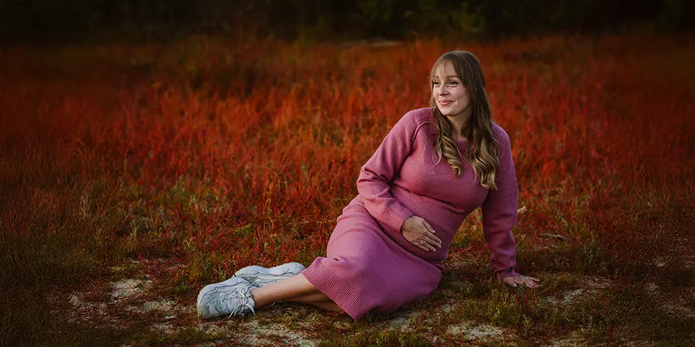 Zwangere vrouw in een roze jurk zit op rood gras en glimlacht rustig, vastgelegd in sfeervolle portretfotografie tijdens een natuurlijke zwangerschapsshoot